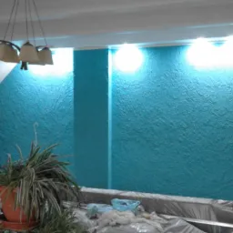 Turkusowa ściana z fakturą, oświetlona od góry, w pomieszczeniu ze skosem. Widoczny fragment lampy i rośliny w doniczce. Ściana zabezpieczona folią malarską.