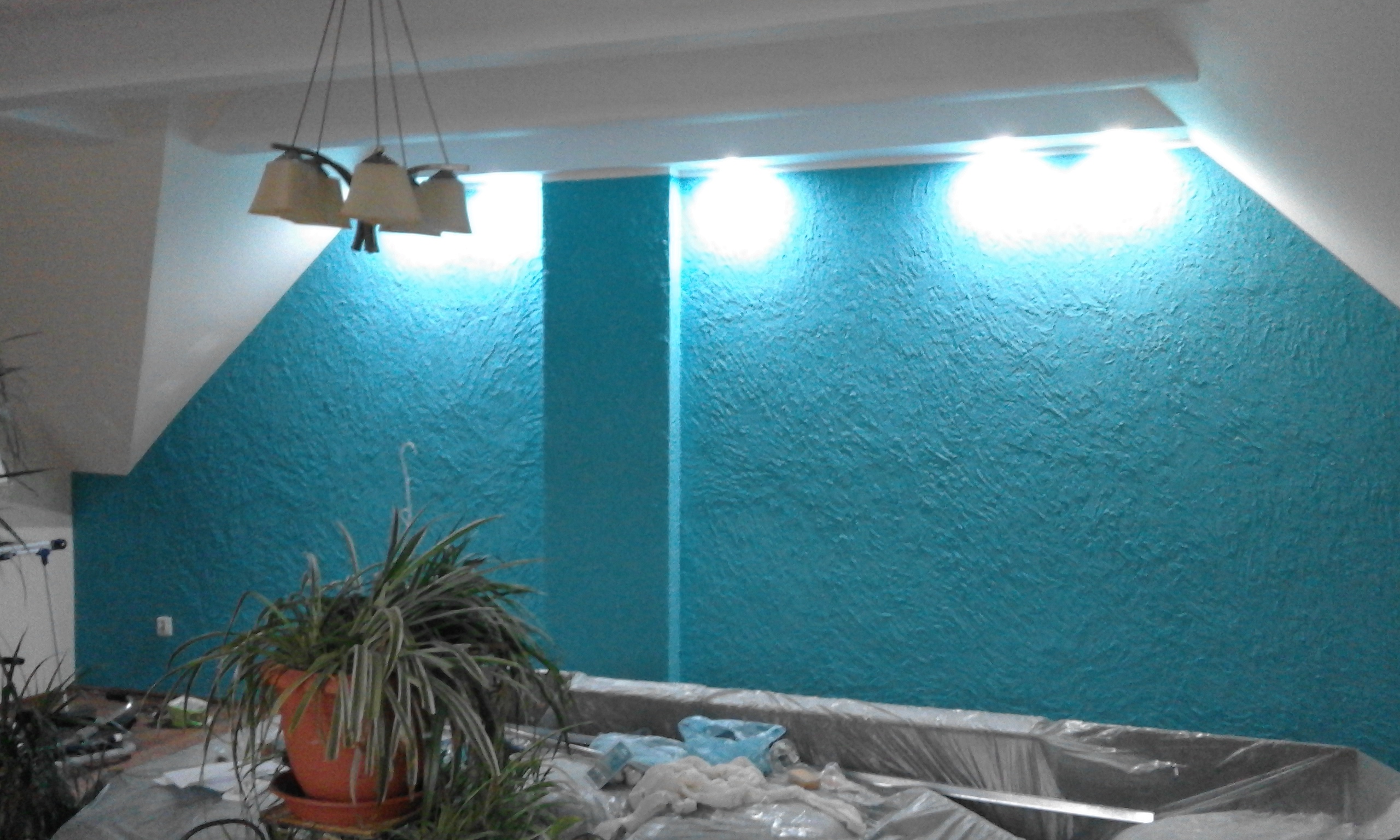 Turkusowa ściana z fakturą, oświetlona od góry, w pomieszczeniu ze skosem. Widoczny fragment lampy i rośliny w doniczce. Ściana zabezpieczona folią malarską.