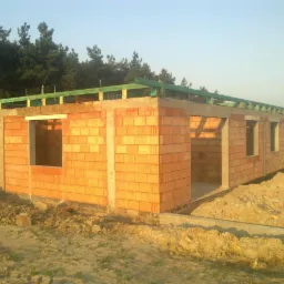 Stan surowy otwarty domu jednorodzinnego z czerwonej cegły, widoczne otwory okienne i drzwiowe, drewniana konstrukcja dachu, tło z zielonymi drzewami.