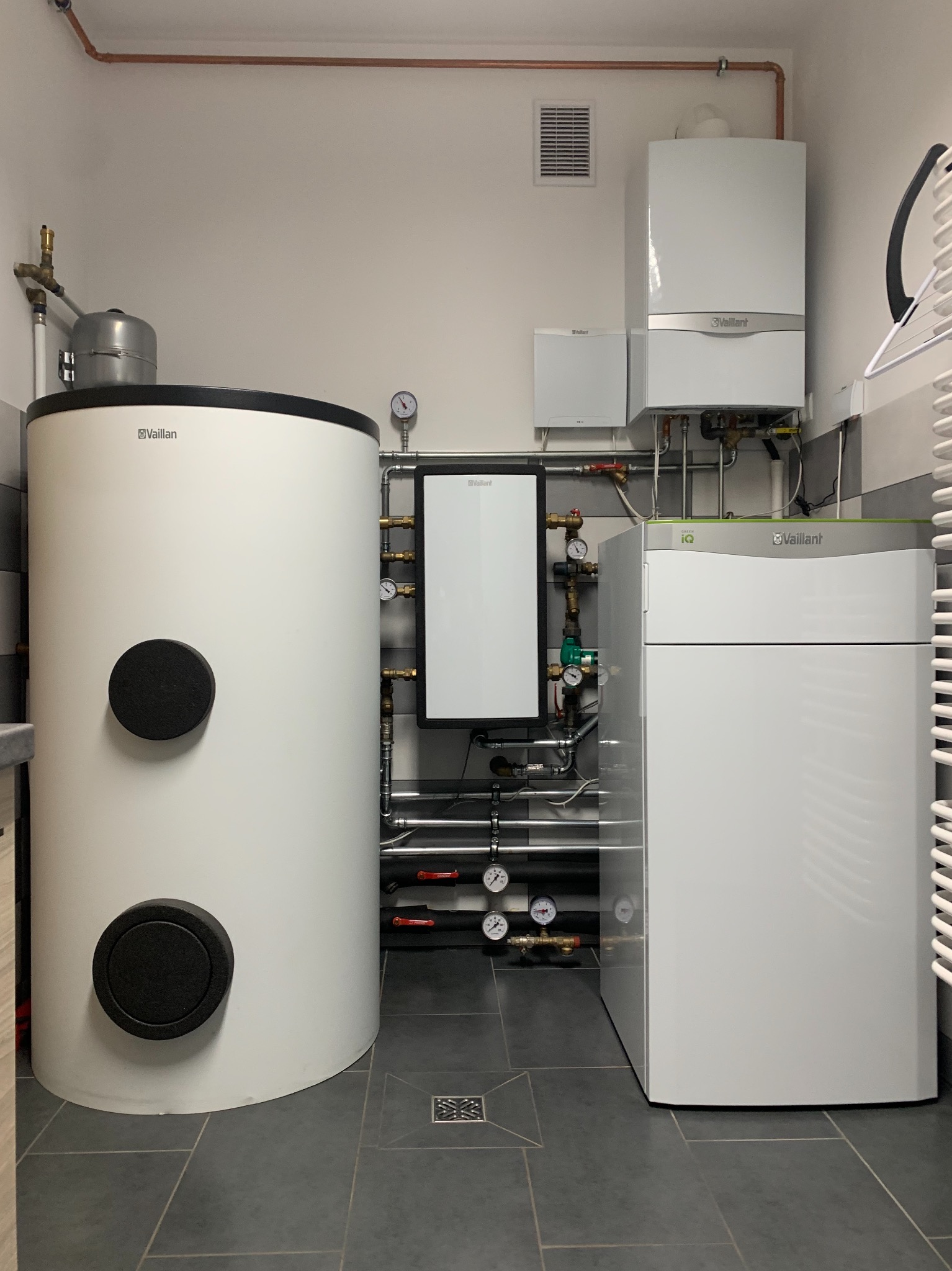 Kompletna instalacja kotłowni: biały zasobnik ciepłej wody, kondensacyjny kocioł gazowy Vaillant Green iQ oraz wiszący kocioł, z widocznymi rurami i manometrami, na tle szarych płytek podłogowych.