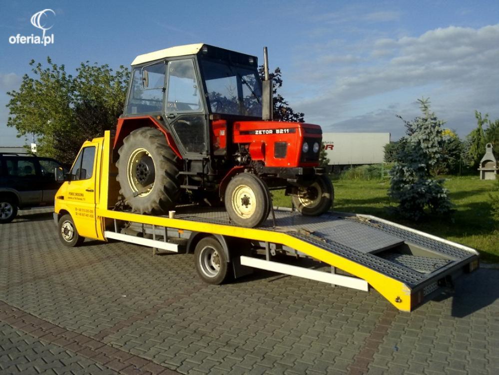 Żółta laweta z czerwonym traktorem Zetor 5211 na platformie, zaparkowana na brukowanym placu w słoneczny dzień. Widoczne drzewa i krzewy w tle.