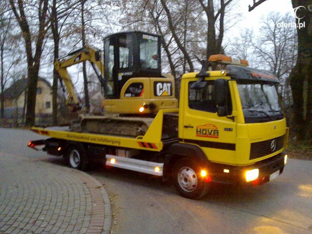 Żółta laweta HOVA przewozi żółtą mini-koparkę CAT na platformie. Widoczne światła awaryjne i ostrzegawcze. Tło stanowią drzewa i budynek.