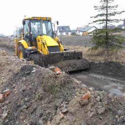 Żółta koparko-ładowarka pracująca na polu, widoczne ślady po wykopie, w tle domy i drzewo iglaste. Na pierwszym planie gruz i ziemia.
