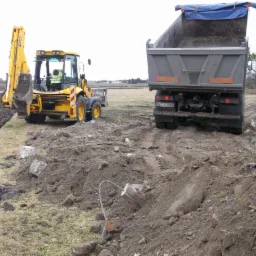 Żółta koparko-ładowarka i wywrotka na placu budowy z gruzem. Wykopany rów w tle. Ślady opon na ziemi, pochmurne niebo. Niwelacja terenu i przygotowanie pod budowę.