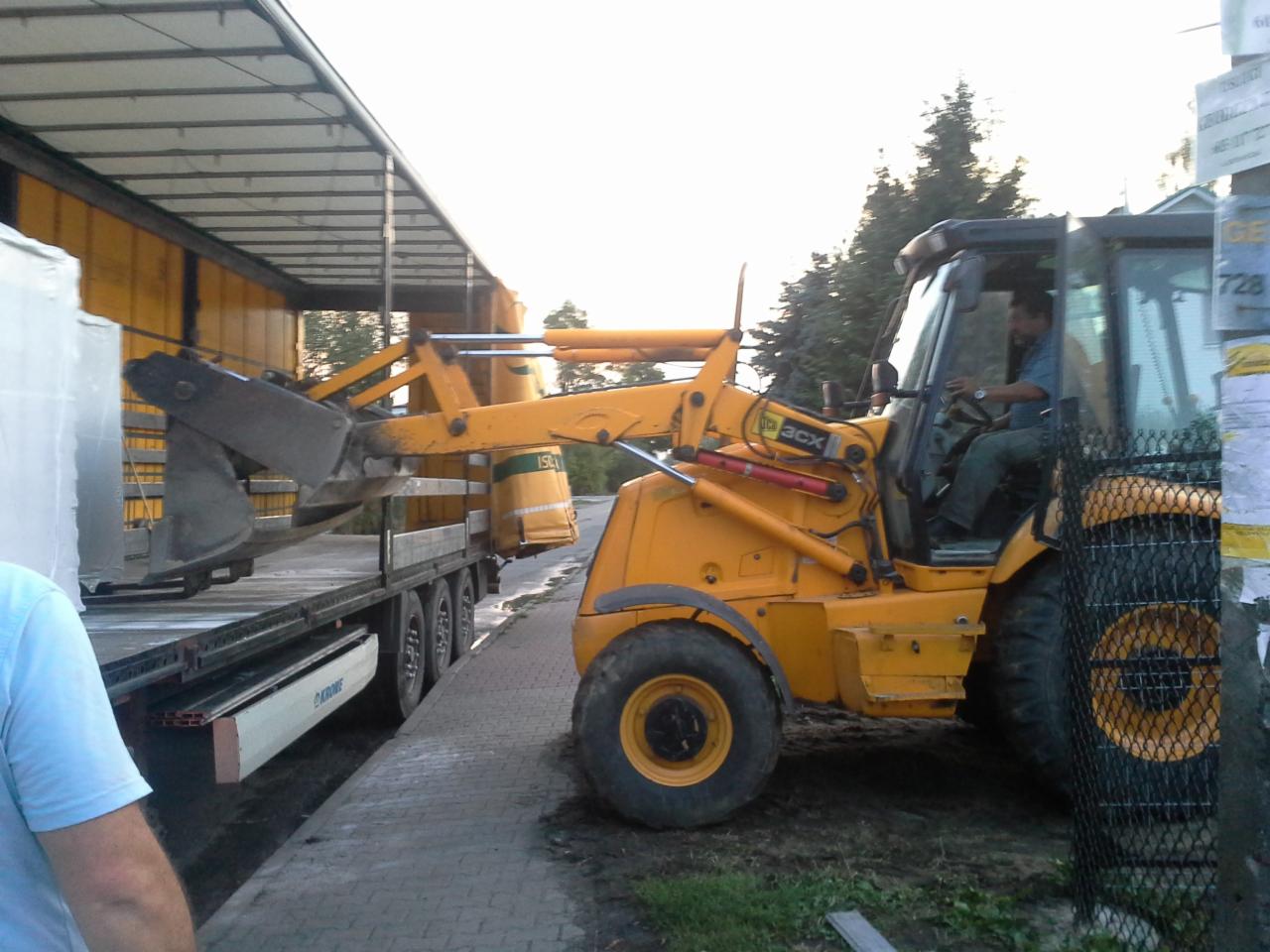 Żółta koparko-ładowarka JCB 3CX załadowuje białe, opakowane materiały na naczepę ciężarówki. Operator w kabinie. Widoczne drzewa w tle, częściowo zasłonięte ogłoszeniami.