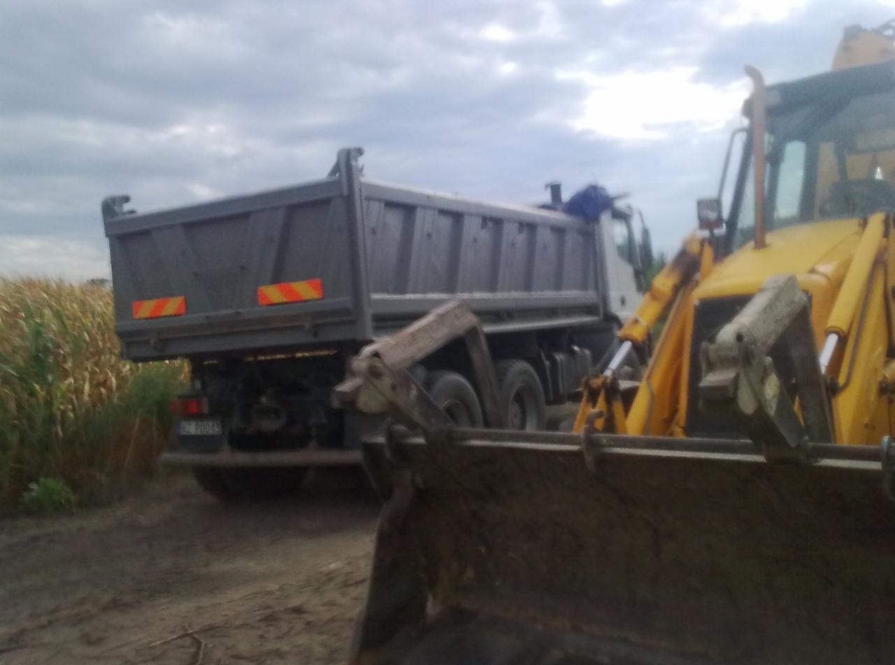 Żółta koparko-ładowarka i wywrotka na tle pola w Baranowie. Sprzęt budowlany gotowy do pracy, pochmurne niebo w tle. Widok z boku, lekko od dołu.