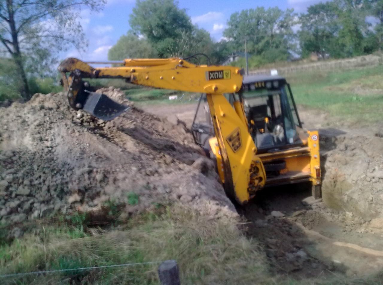 Żółta koparko-ładowarka JCB w trakcie wykopu rowu w ziemi na tle zieleni i błękitnego nieba. Widoczny fragment kabiny operatora i łyżka koparki. Prace ziemne.