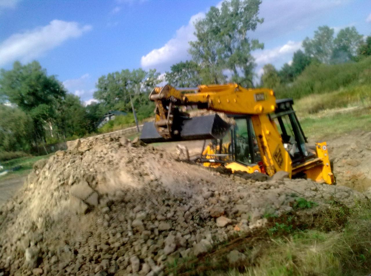Żółta koparko-ładowarka JCB w trakcie prac ziemnych, częściowo zagłębiona w wykopie, z hałdą ziemi przed łyżką na tle zieleni i błękitnego nieba.