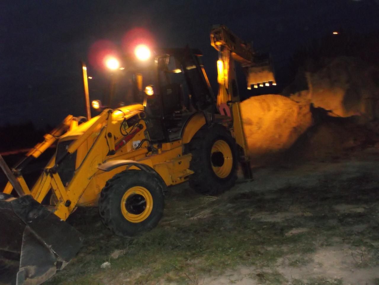 Żółta koparko-ładowarka JCB przy pracy nocą, oświetlona reflektorami, nabiera ziemię z hałdy. Widoczny fragment terenu i ciemne tło.