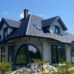 Dom jednorodzinny w trakcie budowy, z czarnym dachem z dachówki modułowej i oryginalnymi oknami w kształcie łuku, widoczne pozostałości materiałów budowlanych na trawniku.