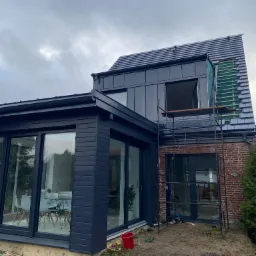 Nowoczesny dom z ciemną elewacją i fragmentem dachu w trakcie krycia, widoczne rusztowanie i przygotowane dachówki.