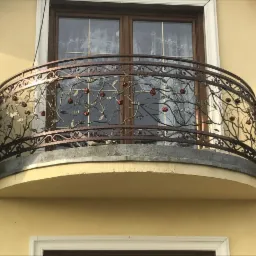 Kuta balustrada balkonowa z motywem różanym, wykonana w Lubsku, na tle okna z firankami.