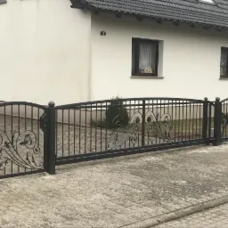 Przesuwna brama wjazdowa z metalu, z dekoracyjnymi elementami roślinnymi, zamontowana przed domem z jasną elewacją i ciemną dachówką.