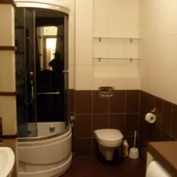 Wykończona łazienka z brązowo-białymi płytkami, narożną kabiną prysznicową i podwieszaną toaletą. Widoczne szklane półki i fragment pralki.