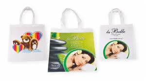 Trzy białe torby bawełniane z różnymi nadrukami: miś z sercami, kobieta z kamieniami do masażu i logo 'La Belle' z portretem kobiety.