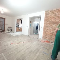 Dawid - Wnętrze w trakcie remontu: ułożona podłoga z płytek imitujących drewno, fragment ściany z cegły, widoczne kable i narzędzia, w tle pomieszczenia z przygotowanymi powierzchniami do malowania.