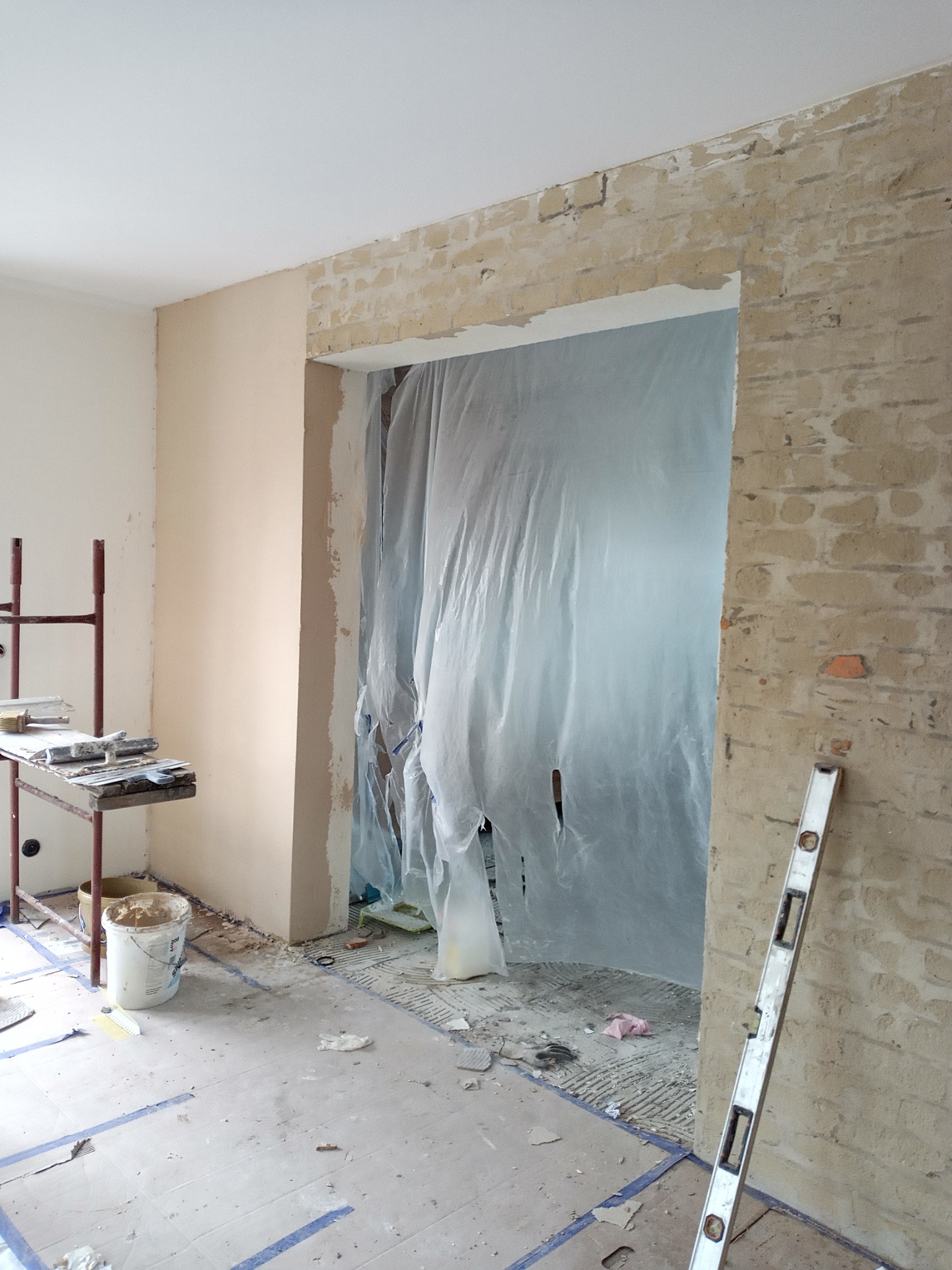 Wnętrze w trakcie prac: ściana częściowo otynkowana, zabezpieczone folią przejście, poziomica oparta o ścianę, widoczne narzędzia i resztki materiałów budowlanych na podłodze.