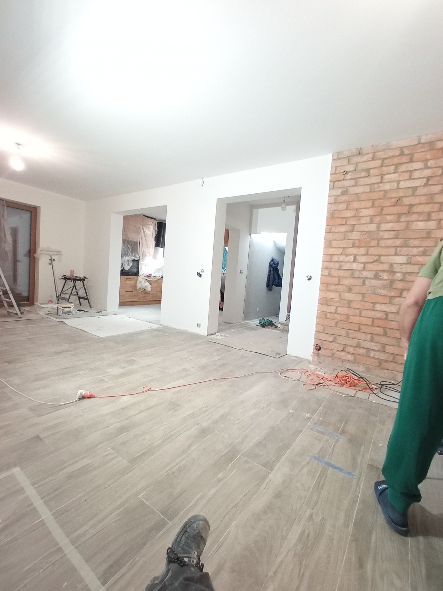 Wnętrze w trakcie remontu: ułożona podłoga z płytek imitujących drewno, fragment ściany z cegły, widoczne kable i narzędzia, w tle pomieszczenia z przygotowanymi powierzchniami do malowania.