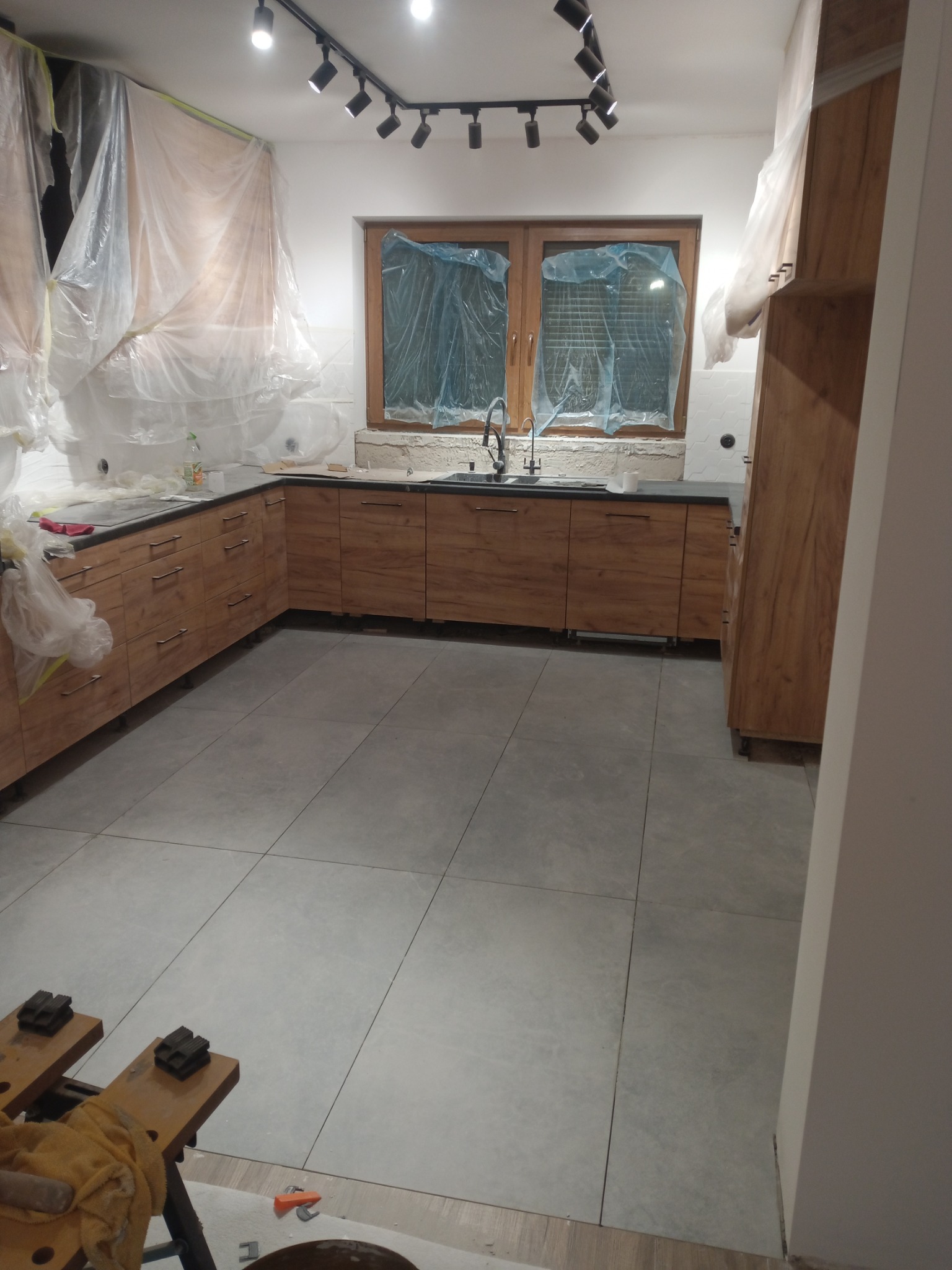 Kuchnia w trakcie prac wykończeniowych: zabezpieczone okna folią, nowa zabudowa meblowa z drewnianymi frontami, szare płytki podłogowe, widoczne narzędzia na podłodze.