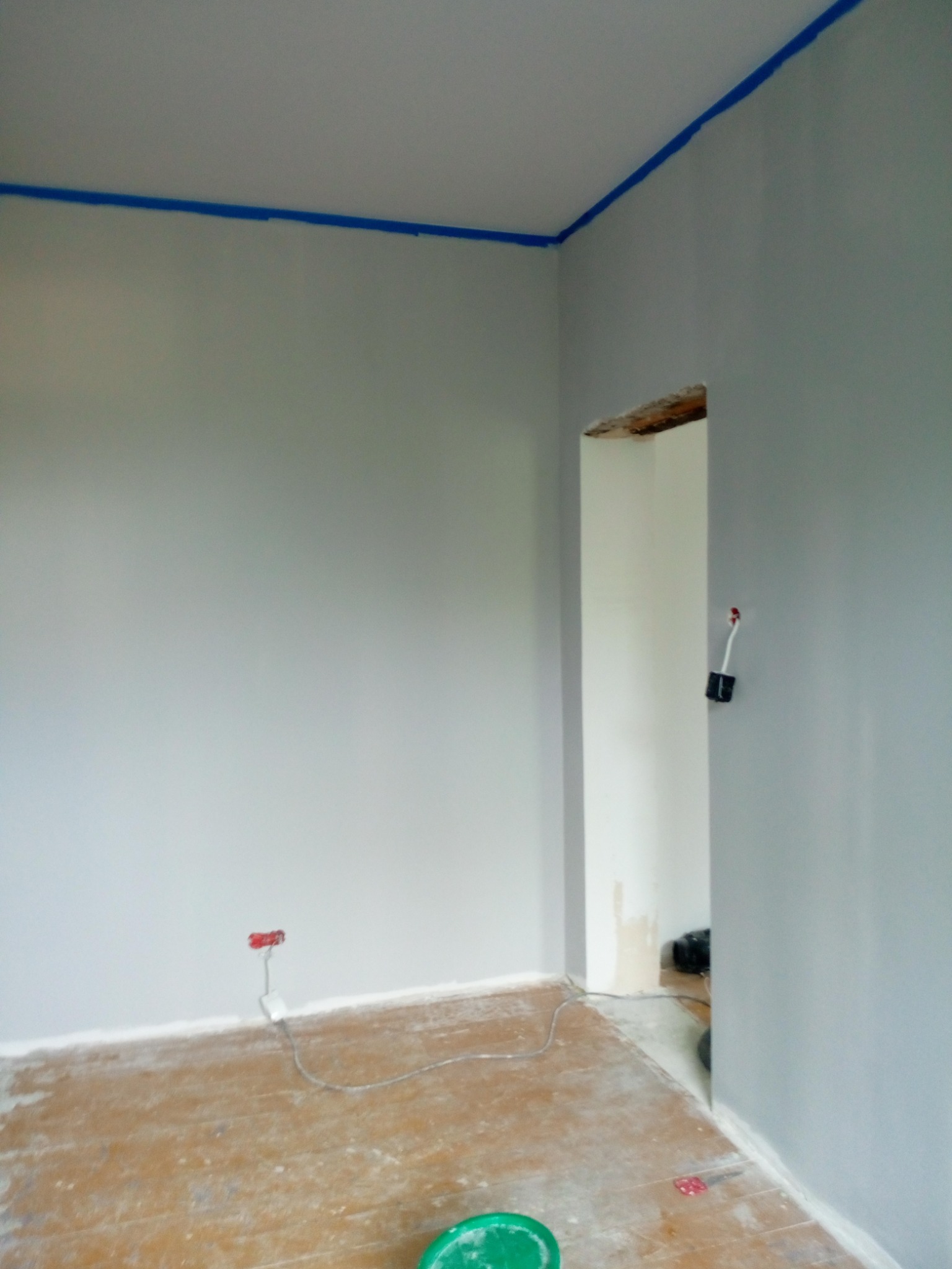 Pokój w trakcie malowania na szaro, z niebieską taśmą malarską przy suficie, widoczne gniazdka elektryczne, podłoga częściowo zakryta folią malarską, ślady farby na podłodze i ścianach.
