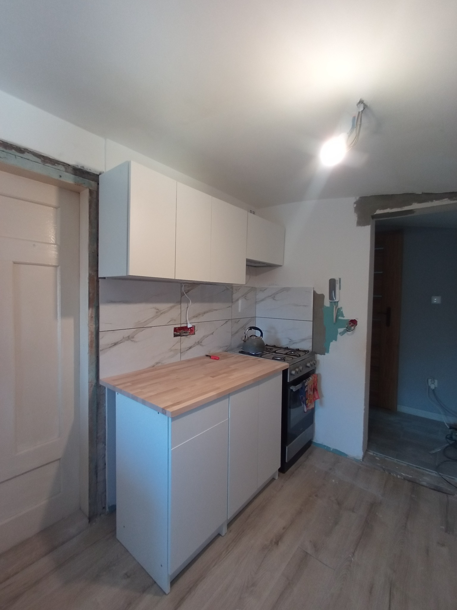 Fragment kuchni w trakcie prac: białe szafki wiszące i stojące z drewnianym blatem, marmurowe płytki na ścianie, widoczne niedokończone gniazdko elektryczne i fragment podłogi z paneli.