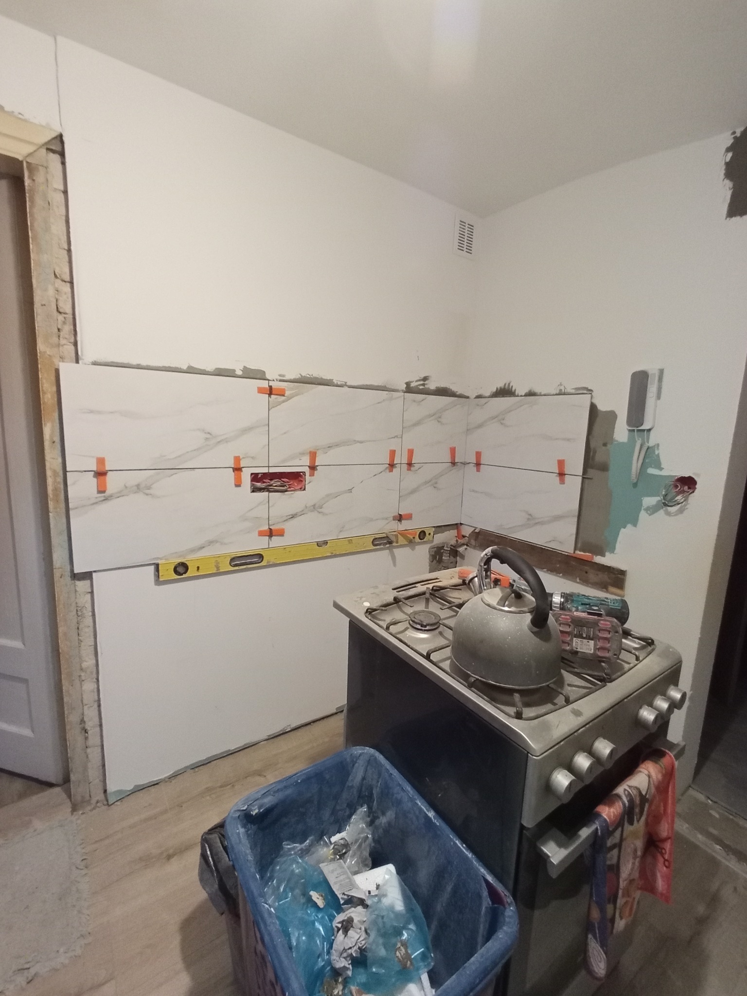 Ściana w trakcie układania płytek imitujących marmur, z pomarańczowymi krzyżykami dystansowymi, poziomica i fragment kuchenki z czajnikiem w tle.