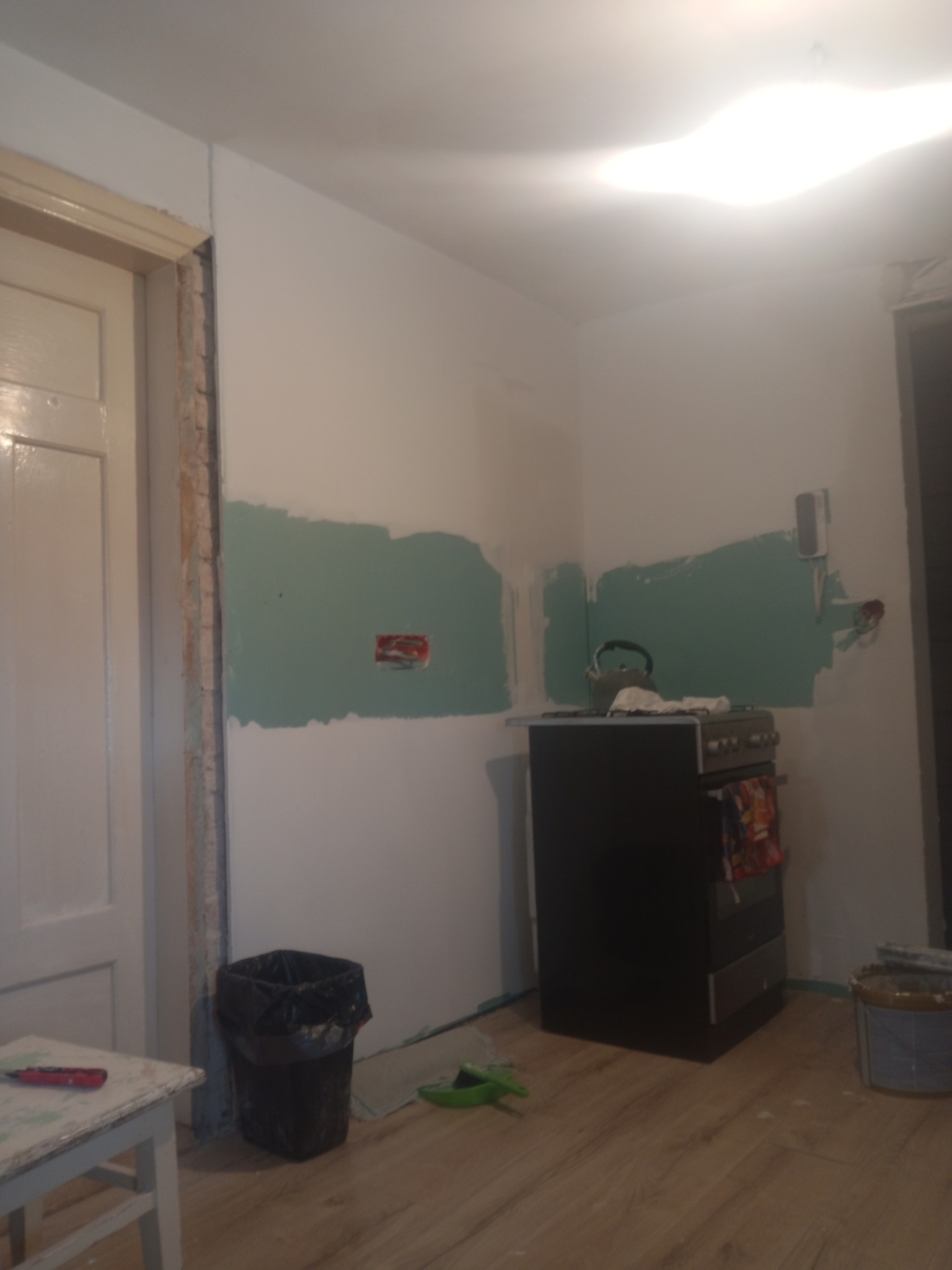 Fragment kuchni w trakcie prac przygotowawczych do malowania: ściany z nałożonym podkładem w kolorze zielonym, widoczny stary piec, śmieci, oraz częściowo zabezpieczone futryny drzwi.