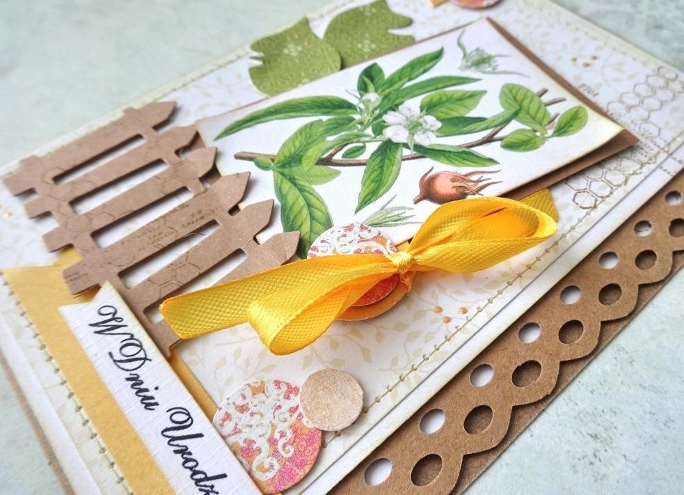 Ręcznie robiona kartka urodzinowa z motywami roślinnymi, żółtą wstążką i tekturowym płotkiem, w ciepłej kolorystyce.