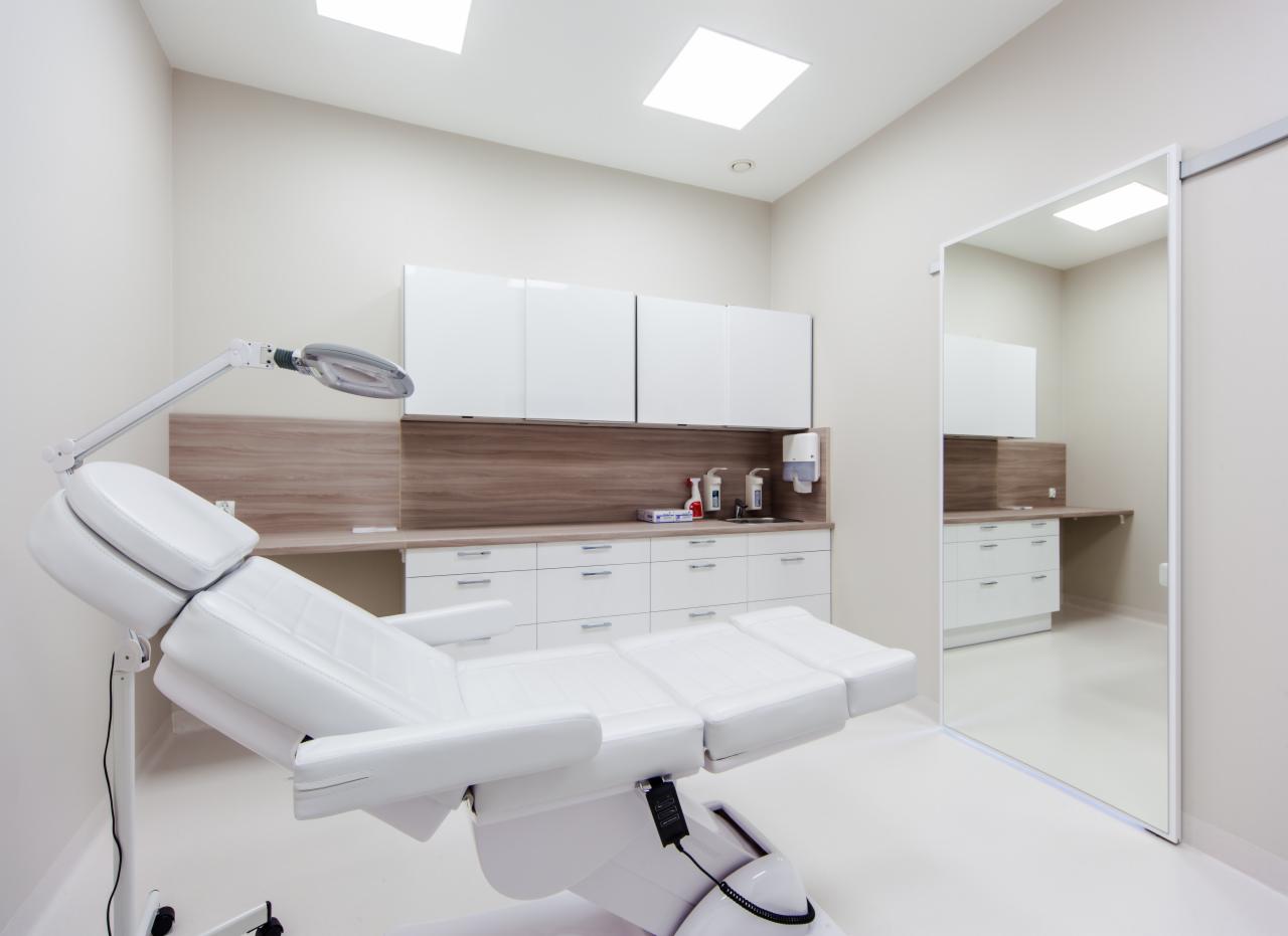 Jasne wnętrze gabinetu lekarskiego z białym fotelem zabiegowym, szafkami i lustrem. Minimalistyczny design, czyste linie i neutralna kolorystyka. Oświetlenie sufitowe.