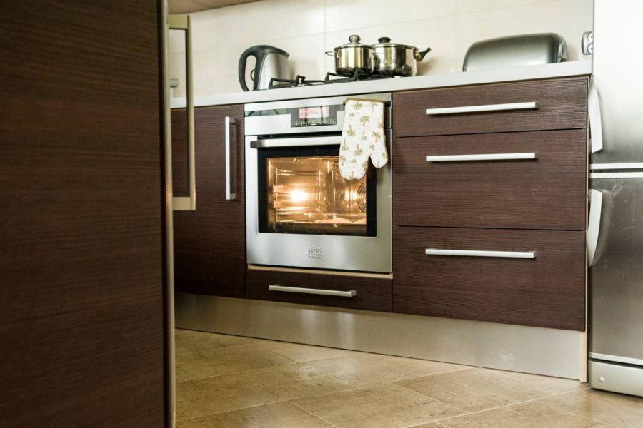 Nowoczesna zabudowa kuchenna z ciemnego drewna z piekarnikiem AEG. Uwagę przykuwają minimalistyczne uchwyty i jasna, ceramiczna podłoga.