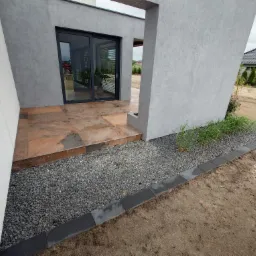 Nowoczesny taras z brązową płytką i szarą elewacją. Mały ogródek z kamykami i obrzeżem z kostki. Widok na dom z przesuwnymi drzwiami.