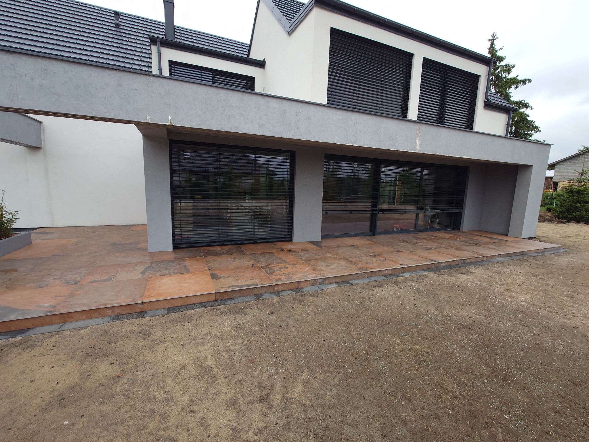 Nowoczesny taras z brązowych płyt, wykończony szarą kostką, przylegający do budynku z szarymi elementami i czarnymi żaluzjami. Widoczny surowy teren wokół.