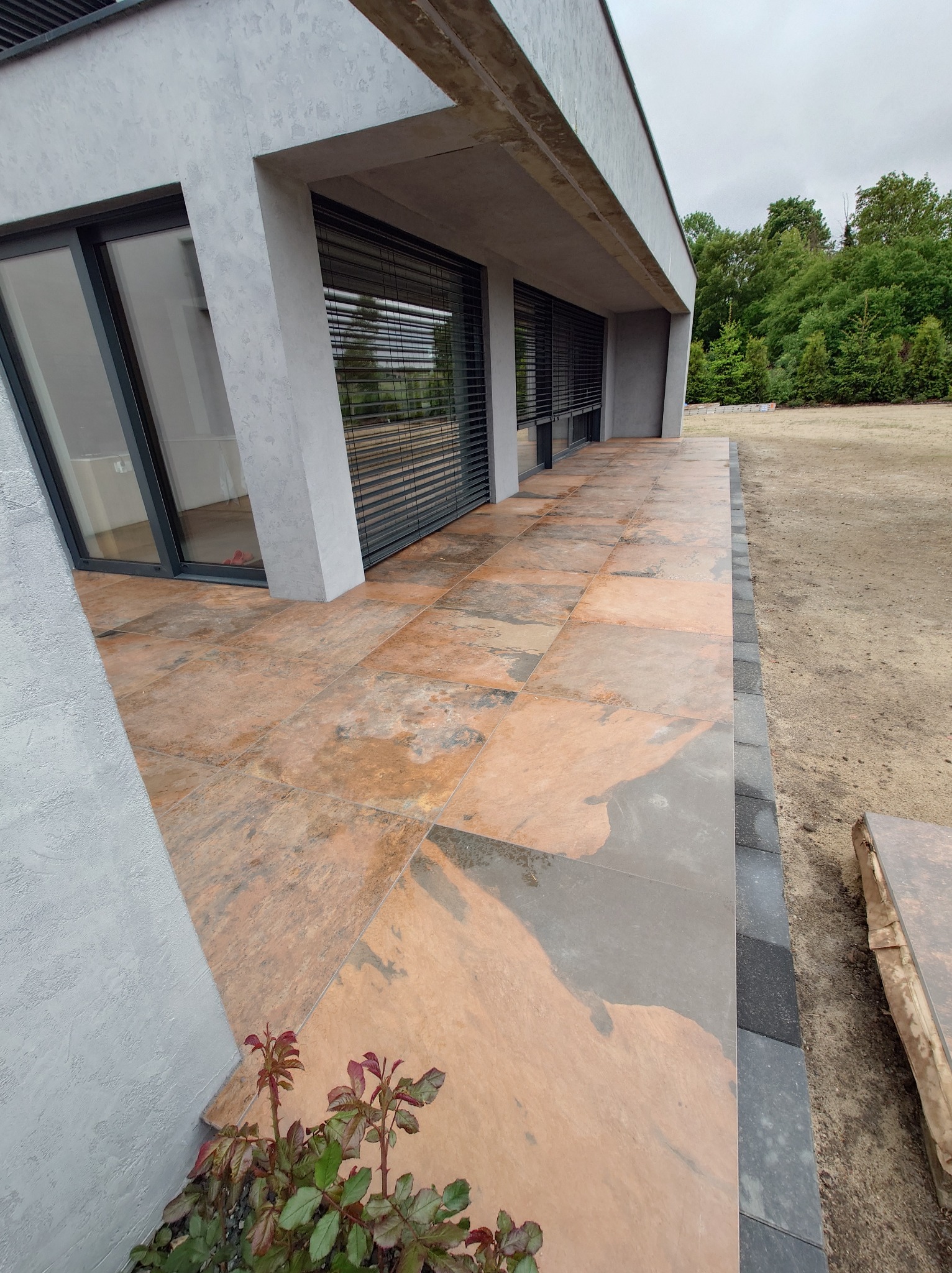 Nowoczesny taras z płyt imitujących kamień, przylegający do budynku o minimalistycznej architekturze z szarym tynkiem strukturalnym i dużymi oknami z żaluzjami.