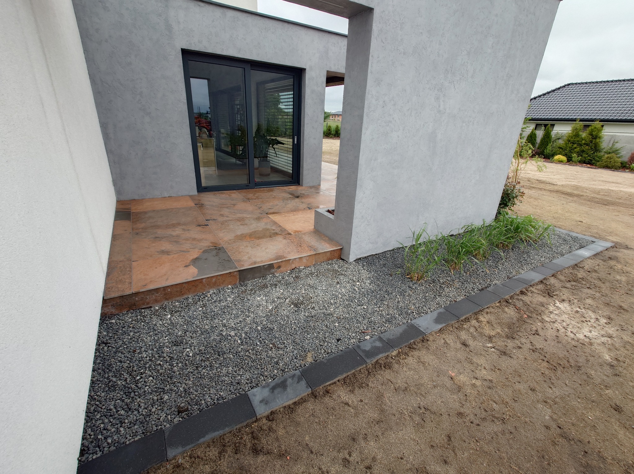 Nowoczesny taras z brązową płytką i szarą elewacją. Mały ogródek z kamykami i obrzeżem z kostki. Widok na dom z przesuwnymi drzwiami.