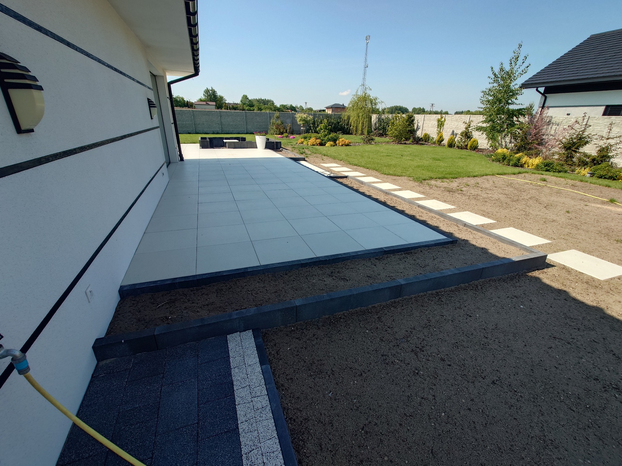 Nowoczesny taras z jasnych płyt, obrzeża z ciemnej kostki, przejście z płyt w trawniku. Widok na zadbany ogród i dom w tle.