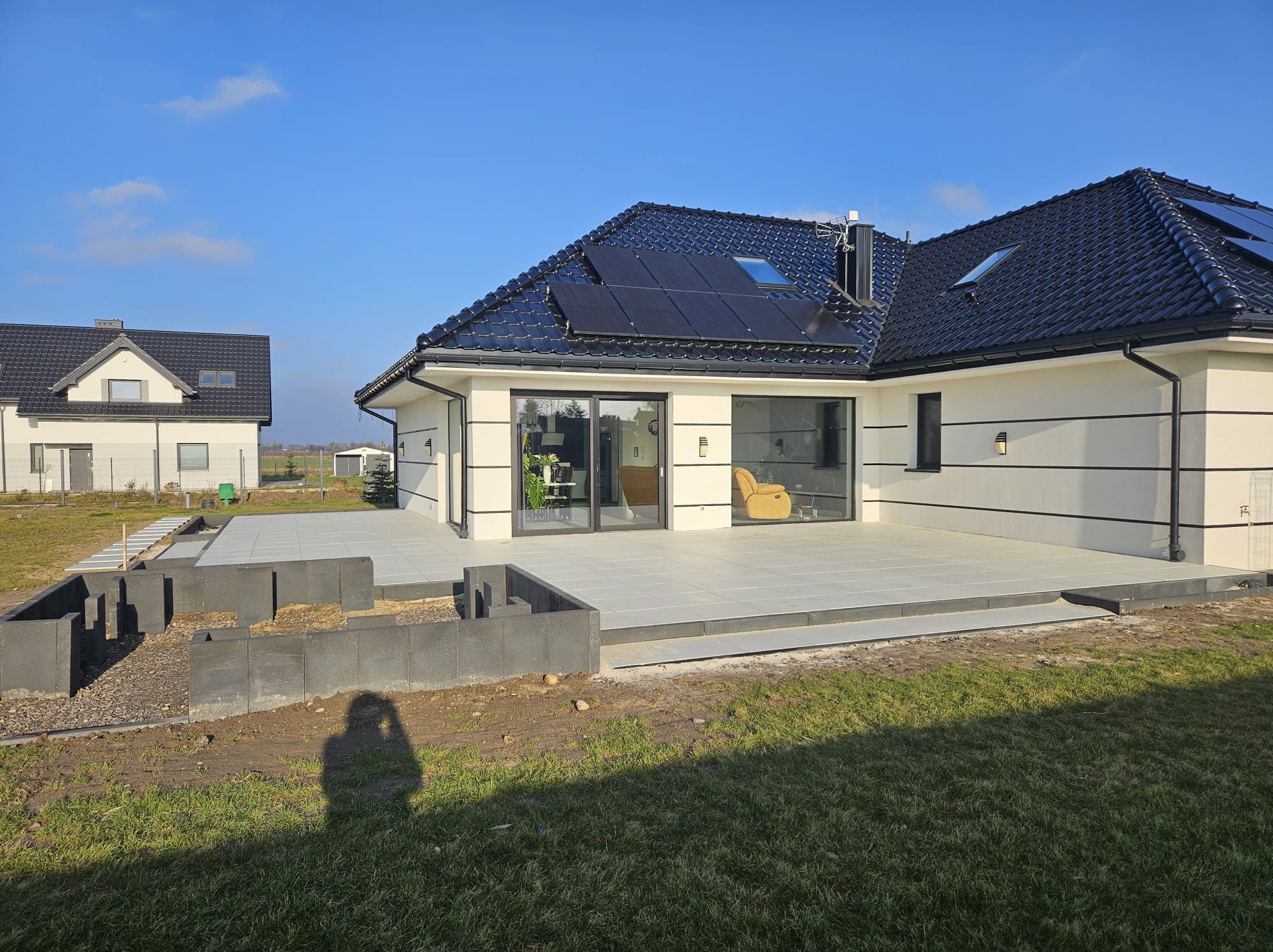 Nowoczesny taras z jasnych płyt, częściowo wykończony, z przygotowanym obrzeżem z szarej kostki. W tle dom z panelami słonecznymi na dachu i zielony trawnik.