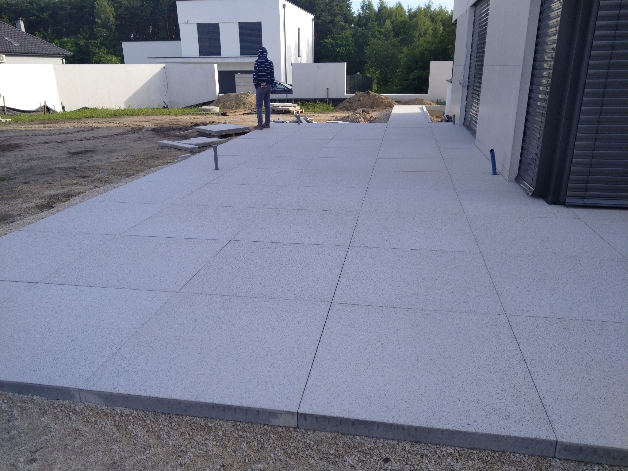 Wykończony taras z jasnoszarych płyt betonowych o drobnej fakturze, obrzeża z szarego betonu, w tle nowoczesny dom z białymi ścianami i szarymi roletami, widoczna osoba w bluzie z kapturem.