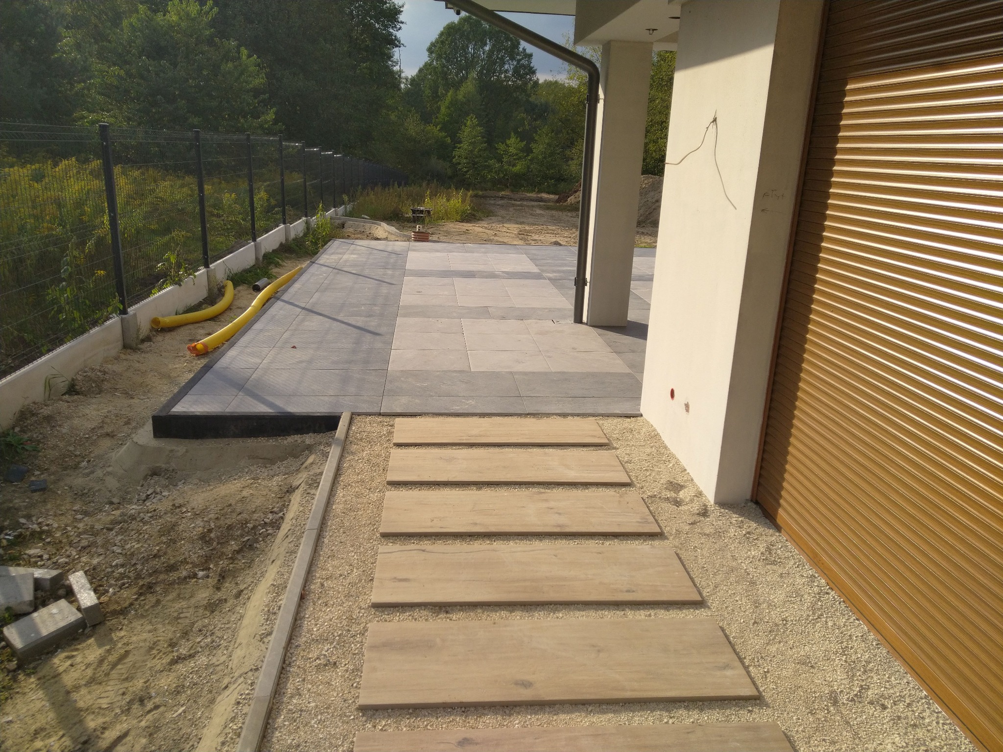 Taras z szarej kostki brukowej i ścieżka z drewnopodobnych płyt na żwirowym podłożu, widoczne przygotowania terenu pod ogród.