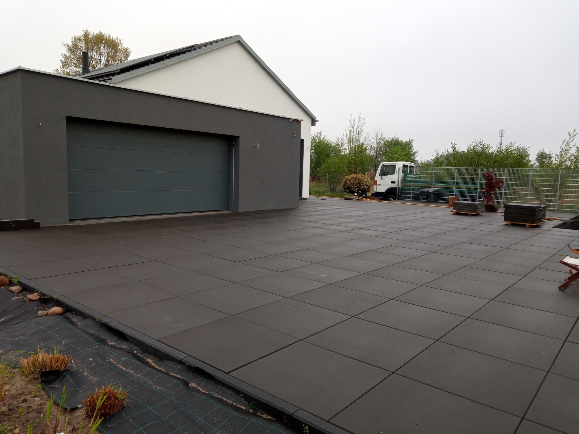 Nowoczesny podjazd z ciemnej kostki brukowej ułożonej przed garażem w szarym kolorze, w tle widoczny biały budynek i samochód dostawczy.