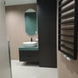 Nowoczesna łazienka z owalnym lustrem, umywalką nablatową na zielonym blacie, czarnymi szafkami, grzejnikiem drabinkowym i beżowymi płytkami ściennymi z delikatnym wzorem.