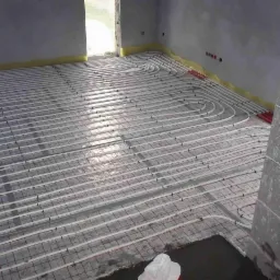 Rozłożone rury grzewcze na folii z siatką, przygotowane do zalania betonem, widok z góry, fragment ściany w surowym stanie.