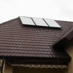 Brązowy dach z trzema kolektorami słonecznymi zamontowanymi na jego powierzchni, widok z dołu na pochmurnym niebie.