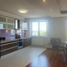 Jasne wnętrze mieszkania w Słupsku: kuchnia z drewnianymi szafkami i czarną lodówką, połączona z jadalnią z widokiem na osiedle przez duże okna.