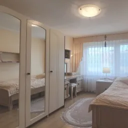 Jasna sypialnia z pojedynczym łóżkiem, toaletką i dużą szafą z lustrami, dywan w kwiaty na podłodze.