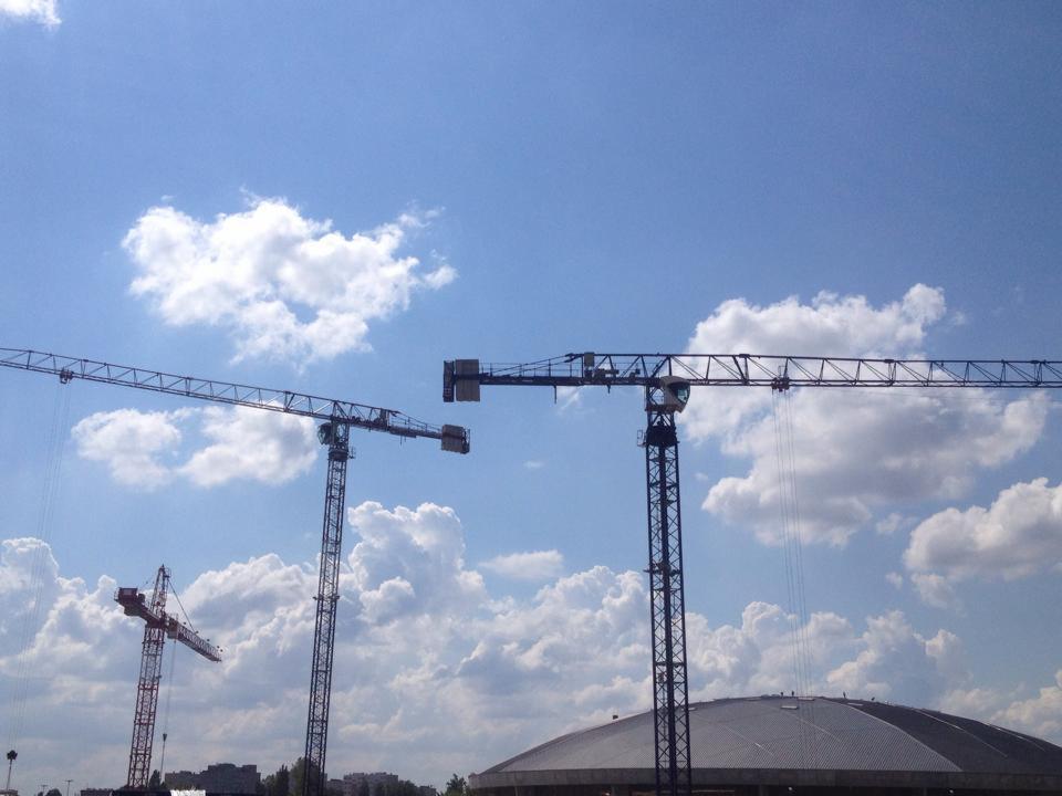 Trzy dźwigi budowlane na tle błękitnego nieba z chmurami, widoczne w oddali budynki i hala sportowa. Ujęcie w słoneczny dzień, z perspektywy żabiej.