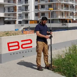 Mężczyzna w brązowych spodniach i granatowej koszulce obsługuje precyzyjny instrument pomiarowy na tle nowego budynku mieszkalnego z oznaczeniem '29G' i betonowego murku z czerwonym logo 'B2...