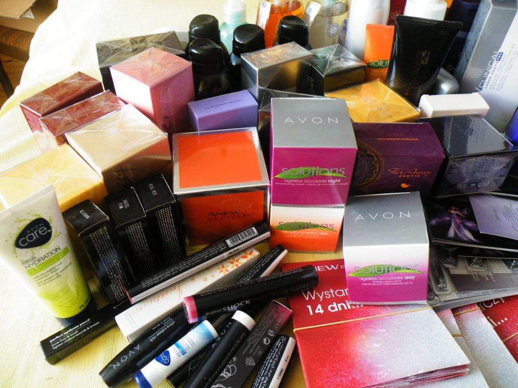 Kolekcja kosmetyków Avon: kremy, szminki, tusze do rzęs, pudry i inne produkty do makijażu i pielęgnacji, ułożone na jasnym tle. Różnorodność opakowań i kolorów.