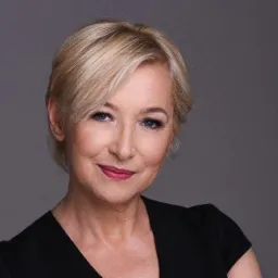 Portret kobiety z krótkimi blond włosami, delikatny uśmiech, makijaż w odcieniach różu, na szarym tle.