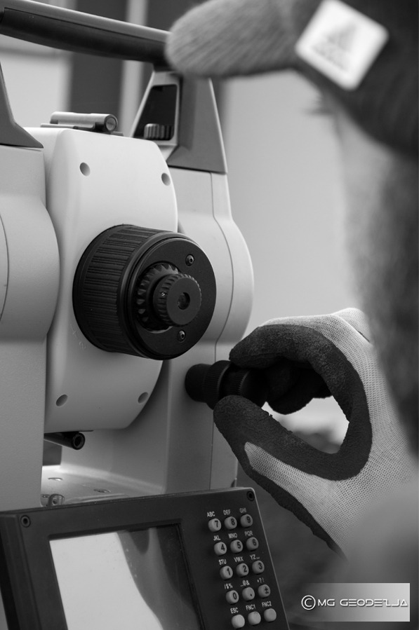 Czarno-białe zbliżenie na teodolit, dłoń w rękawicy reguluje ustawienia, widoczny panel sterowania z klawiaturą numeryczną.