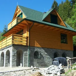 Dwukondygnacyjny dom z bali z balkonem, zielonym dachem i kamienną podmurówką z łukami, na tle lasu i błękitnego nieba. Widoczne materiały budowlane.